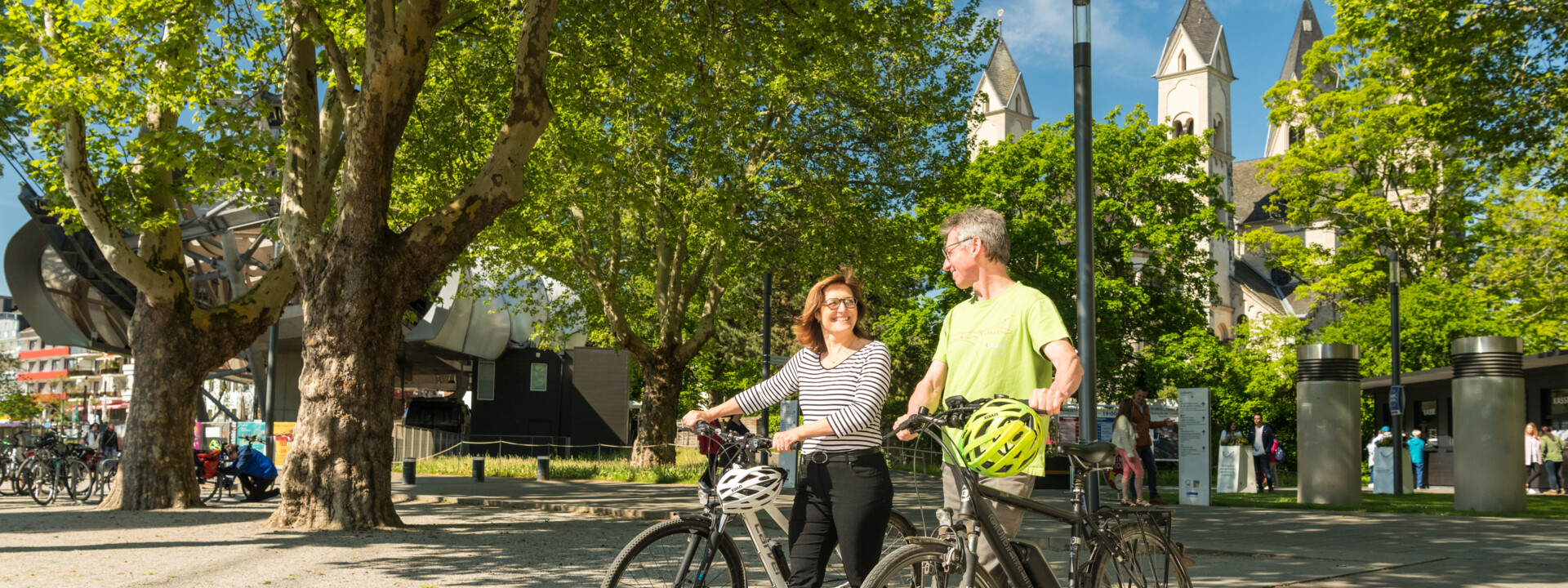 Pärchen mit E-Bikes lächeln. Seilbahn-Talstation und Basilika St. Kastor sind im Hintergrund zu sehen. © Koblenz-Touristik GmbH, Dominik Ketz Pärchen mit E-Bikes lächeln. Seilbahn-Talstation und Basilika St. Kastor sind im Hintergrund zu sehen. © Koblenz-Touristik GmbH, Dominik Ketz