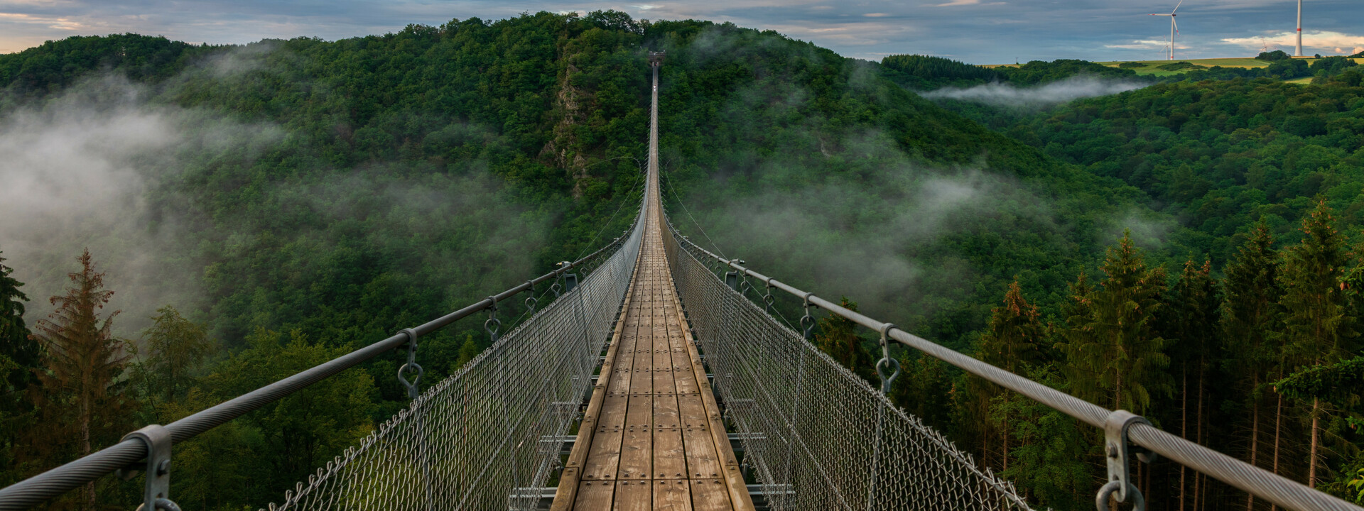 Hängebrücke Geierlay © Bernhard - stock.adobe.com Blick auf eine lange Hängeseilbrücke, die sich über ein tiefes bewaldetes Tal erhebt, im Hintergrund bewölkter Himmel und Nebelschwaden im Tal  © Bernhard - stock.adobe.com