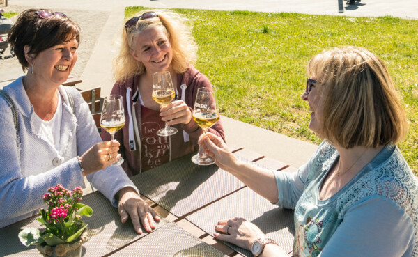 Drei Damen sitzen an einem Tisch, lächeln und stoßen mit Weingläsern an, im Hintergrund grüne Wiese und das Preußische Regierungsgebäude © Dominik Ketz Drei Damen sitzen an einem Tisch, lächeln und stoßen mit Weingläsern an, im Hintergrund grüne Wiese und das Preußische Regierungsgebäude © Dominik Ketz