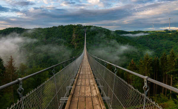 Blick auf eine lange Hängeseilbrücke, die sich über ein tiefes bewaldetes Tal erhebt, im Hintergrund bewölkter Himmel und Nebelschwaden im Tal  © Bernhard - stock.adobe.com Blick auf eine lange Hängeseilbrücke, die sich über ein tiefes bewaldetes Tal erhebt, im Hintergrund bewölkter Himmel und Nebelschwaden im Tal  © Bernhard - stock.adobe.com