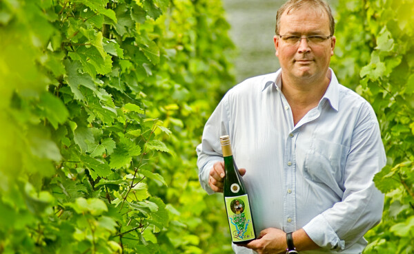 Winzer Rainer Kollman steht mit einer Flasche Wein in seinem Weinberg © Fotografenklasse der Julius-Wegeler-Schule Winzer Rainer Kollman steht mit einer Flasche Wein in seinem Weinberg © Fotografenklasse der Julius-Wegeler-Schule
