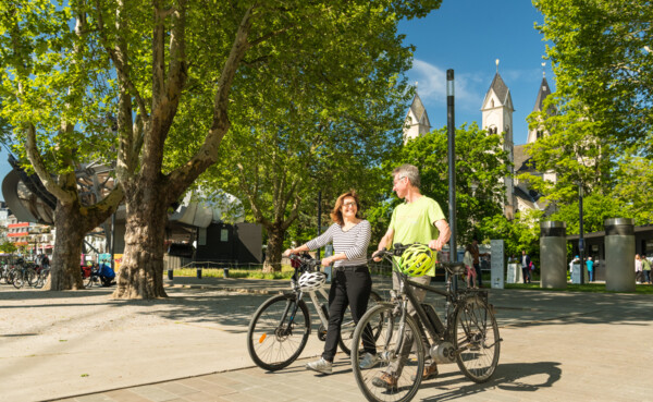 Pärchen E-Bikes Fahrrad Koblenz © Koblenz-Touristik GmbH, Dominik Ketz Pärchen mit E-Bikes lächeln. Seilbahn-Talstation und Basilika St. Kastor sind im Hintergrund zu sehen. © Koblenz-Touristik GmbH, Dominik Ketz