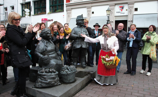 Stadtführung Heilije Hexe Huckeweiwer © Koblenz-Touristik GmbH, Juraschek Verkleidete Stadtführerin mit Korb und Gemüse lacht mit einer Führungsgruppe vor Figuren auf dem Münzplatz © Koblenz-Touristik GmbH, Juraschek