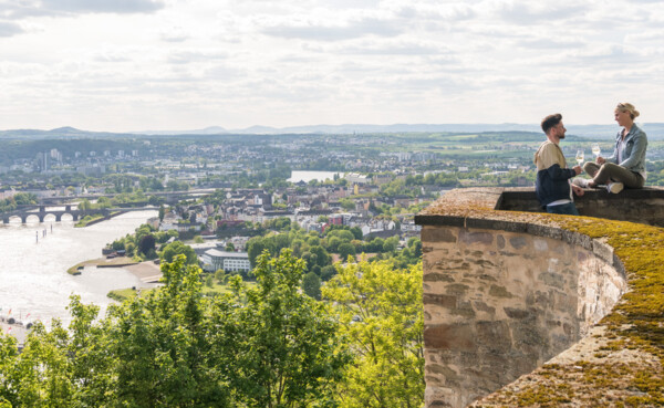 Pärchen sitzt auf der Mauer der Festung Ehrenbreitstein und hält Weingläser mit Stadt Koblenz im Hintergrund © Koblenz-Touristik GmbH, Dominik Ketz