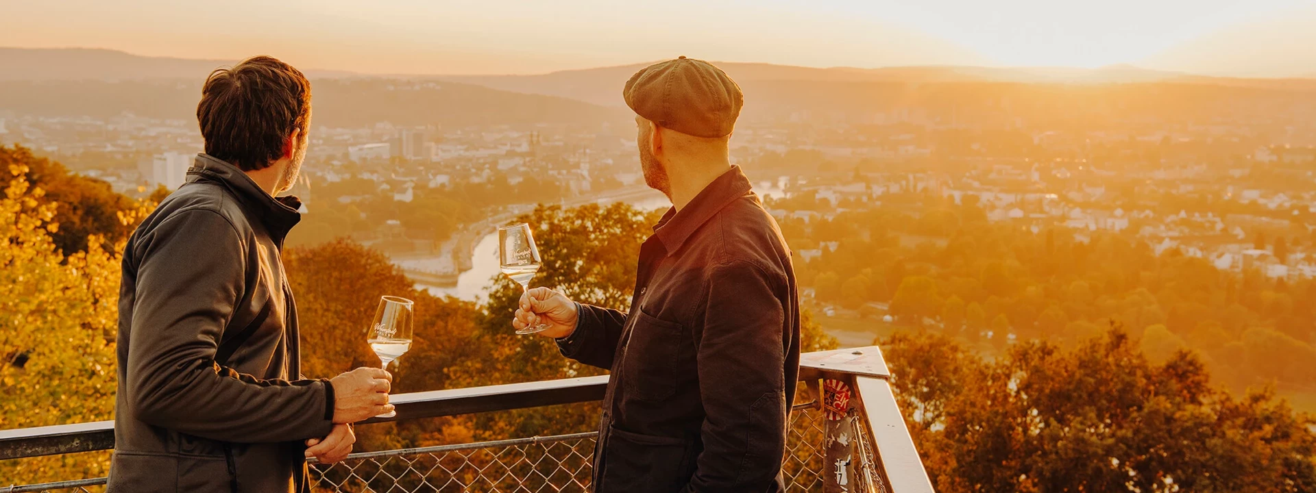 Zwei M&auml;nner von der Seite halten Weingl&auml;ser und blicken auf die Stadt Koblenz beim Sonnenuntergang &copy; Koblenz-Touristik GmbH, Philip Bruederle