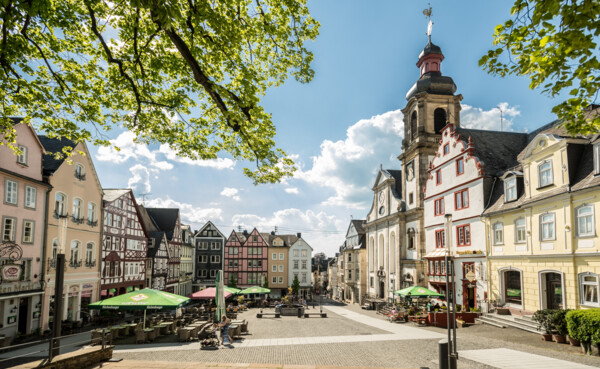 Hachenburger Marktplatz mit Tischen und Sonnenschirmen vor Restaurants, umgeben von farbenfrohen Fachwerkhäusern und einer Kirche, im Vordergrund Äste, im Hintergrund blauer Himmel © Tourist-Info Hachenburger Westerwald, Dominik Ketz Hachenburger Marktplatz mit Tischen und Sonnenschirmen vor Restaurants, umgeben von farbenfrohen Fachwerkhäusern und einer Kirche, im Vordergrund Äste, im Hintergrund blauer Himmel © Tourist-Info Hachenburger Westerwald, Dominik Ketz