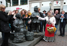 Stadtführung Heilije Hexe Huckeweiwer © Koblenz-Touristik GmbH, Juraschek Verkleidete Stadtführerin mit Korb und Gemüse lacht mit einer Führungsgruppe vor Figuren auf dem Münzplatz © Koblenz-Touristik GmbH, Juraschek