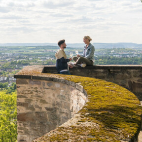 Pärchen Mauer Festung Ehrenbreitstein Koblenz © Koblenz-Touristik GmbH, Dominik Ketz Pärchen sitzt auf der Mauer der Festung Ehrenbreitstein und hält Weingläser mit Stadt Koblenz im Hintergrund © Koblenz-Touristik GmbH, Dominik Ketz