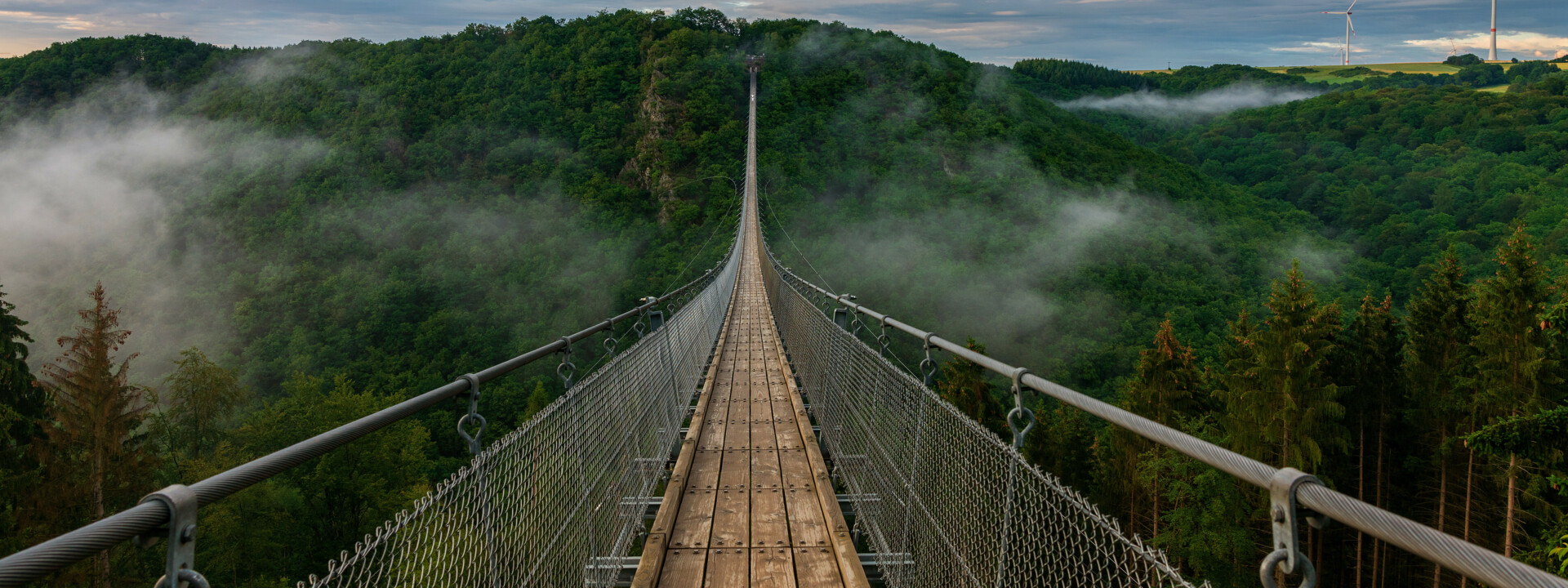 Hängebrücke Geierlay © Bernhard - stock.adobe.com Blick auf eine lange Hängeseilbrücke, die sich über ein tiefes bewaldetes Tal erhebt, im Hintergrund bewölkter Himmel und Nebelschwaden im Tal  © Bernhard - stock.adobe.com