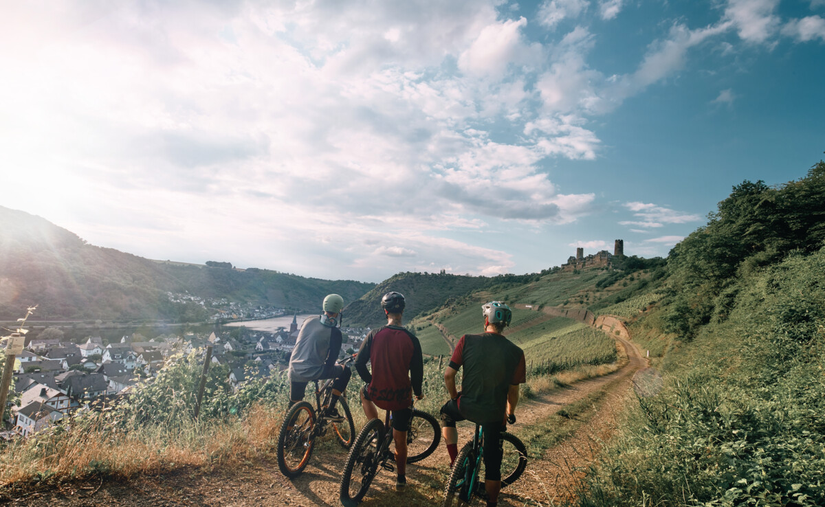 Freundesgruppe auf Mountainbikes auf einem Radweg neben der Mosel mit Burg Thurant im Hintergrund © Philip Bruederle