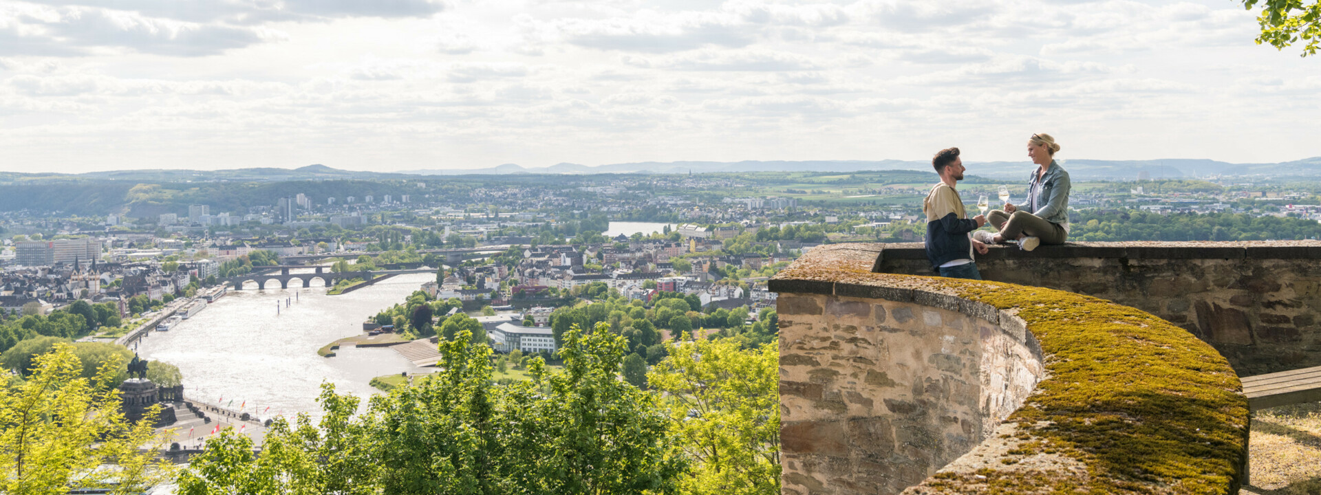 Pärchen Mauer Festung Ehrenbreitstein Koblenz © Koblenz-Touristik GmbH, Dominik Ketz Pärchen sitzt auf der Mauer der Festung Ehrenbreitstein und hält Weingläser mit Stadt Koblenz im Hintergrund © Koblenz-Touristik GmbH, Dominik Ketz